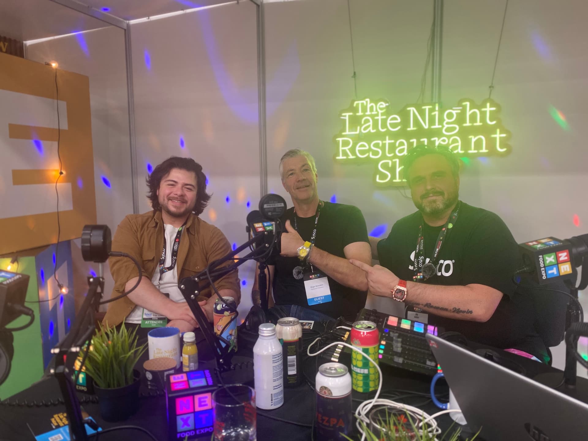 Jay Ashton and Domenic Pedulla recording with a guest under the neon The Late Night Restaurant Podcast sign at NEXT Food Expo
